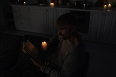 Mutfaktaki elektrik kesintisi sırasında yanan mumlarla kitap okuyan adamın yüksek açılı görüntüsü 