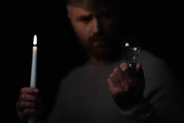 在用黑色隔离的能源停电时 拿着灯泡和点燃的蜡烛的模糊男子 — 图库照片