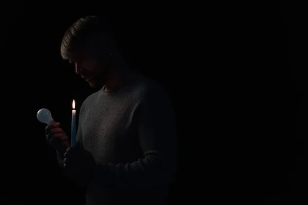 man in darkness holding lit candle and electric bulb isolated on black