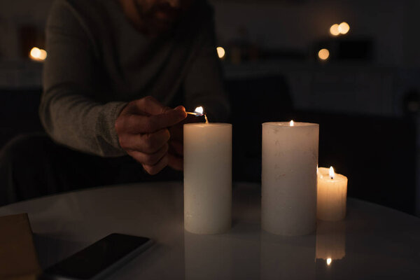 обрезанный вид человека в темноте зажигая свечу с горящей спичкой возле смартфона с пустым экраном