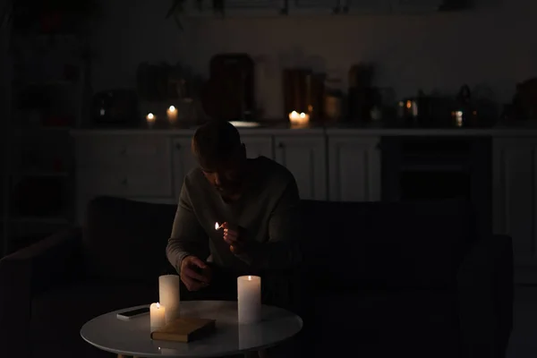 男人坐在黑暗的厨房里 点燃蜡烛 拿着智能手机看书 — 图库照片