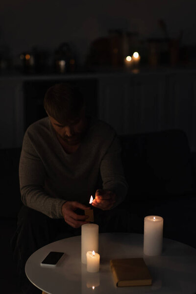 человек, сидящий в темноте и зажигая свечи возле книги и мобильного телефона с пустым экраном