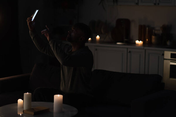 man sitting in darkness near burning candles and catching mobile connection on smartphone