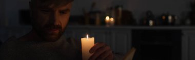 man holding burning candle in dark kitchen during electricity outage, banner clipart