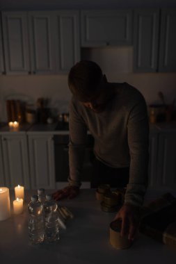 Karanlık mutfakta duran adam konserve yiyeceklerin yanında şişe su ve mumlarla bekliyor.