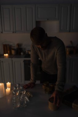 man standing in dark kitchen near stock of canned food with bottled water and candles clipart