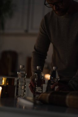 Şişelenmiş suyun yanında gözlüklü bir adam ve mutfakta sıcak battaniyeli konserve yiyecekler.