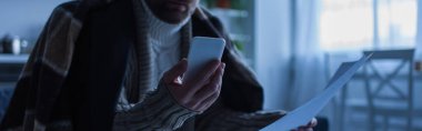 cropped view of man holding smartphone and payment invoices during power blackout, banner clipart