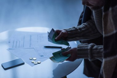 Alacakaranlıkta akıllı telefon ve ödeme faturalarının yanında para sayan adamın kısmi görüntüsü