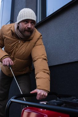 Örgü şapkalı ve ceketli bir adam güç jeneratörünü çalıştırıyor ve dışarı bakıyor.