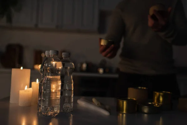 暗いキッチンのクロップドマンに近いキャンドル付きボトル入りの水と缶詰の食べ物の準備 — ストック写真