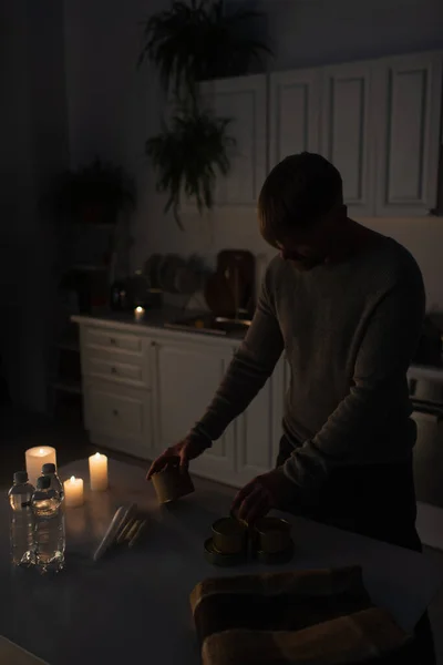 man holding canned food near bottled water and candles in kitchen during power blackout