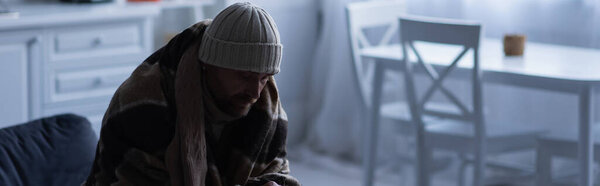 Замороженный человек сидит в теплом одеяле и вязаной шляпе дома в сумерках, баннер