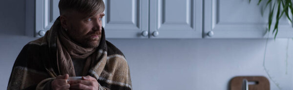 замороженный человек в теплом одеяле, держа чашку горячего чая и глядя на дом в сумерках, баннер