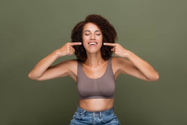 Positive african american woman in top pointing with fingers at mouth isolated on green  clipart