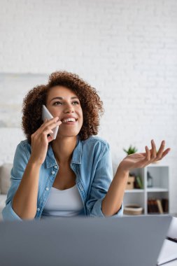 Neşeli Afro-Amerikan Serbest Çalışan akıllı telefonuyla evde bulanık dizüstü bilgisayarın yanında konuşuyor. 
