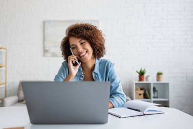 Pozitif Afrikalı Amerikalı serbest çalışan cep telefonuyla konuşuyor ve oturma odasında dizüstü bilgisayara bakıyor. 