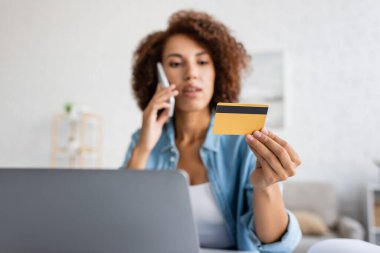 Bulanık Afrikalı Amerikalı kadın akıllı telefonla konuşuyor ve evinde dizüstü bilgisayarının yanında kredi kartı tutuyor. 
