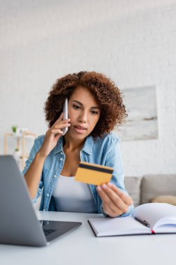 Afro-Amerikalı bir kadın akıllı telefondan konuşuyor ve evinde bulanık bir kredi kartı tutuyor. 