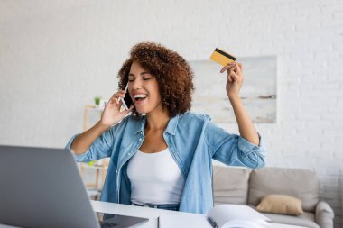 Heyecanlı Afrikalı Amerikalı kadın kredi kartı tutuyor ve evde online alışveriş yaparken akıllı telefondan konuşuyor. 