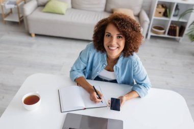 Neşeli Afro-Amerikalı kadın elinde akıllı telefonuyla dizüstü bilgisayarın yanında yazı yazıyor.