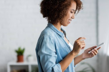 Gülümseyen kıvırcık saçlı, akıllı telefon kullanan Afro-Amerikan kadının yan görüntüsü 