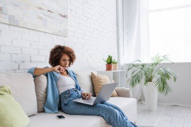 Düşünceli Afrikalı Amerikalı kadın koltukta otururken ve evde çalışırken dizüstü bilgisayar kullanıyor. 