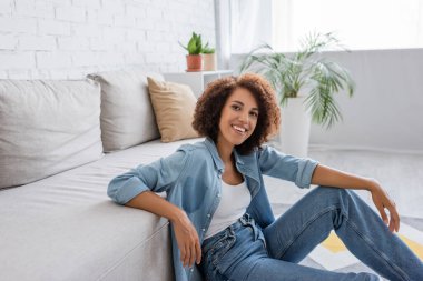 Pozitif Afro-Amerikalı kıvırcık saçlı kadın oturma odasında modern kanepenin yanında oturuyor.