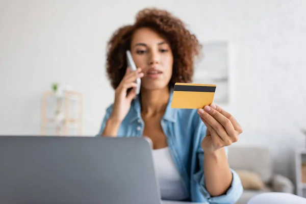 Blurred african american woman talking on smartphone and holding credit card near laptop at home