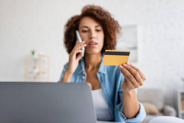Blurred african american woman talking on smartphone and holding credit card near laptop at home 