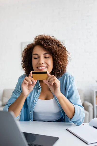 Pleased african american woman holding credit card near blurred laptop and notebook at home