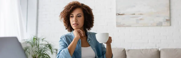 沉思的非洲裔美国女人举着一杯咖啡 看着笔记本电脑 一面横幅 — 图库照片