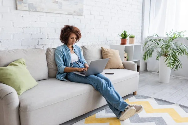 卷曲的非洲裔美国自由职业者坐在沙发上在家工作时使用笔记本电脑 — 图库照片