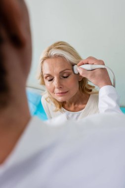 Orta yaşlı kadın gözleri kapalı otururken doktor ön planda kafa ultrasonu yapıyor.