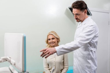 Doktor, orta yaşlı gülümseyen bir kadının yanındaki ultrason cihazını işaret ediyor.