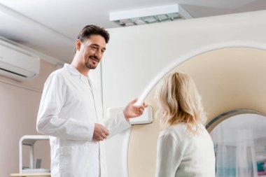 Gülümseyen doktor, orta yaşlı sarışın bir kadının yanında bilgisayarlı tomografi tarayıcısını işaret ediyor.