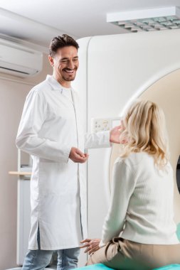 Beyaz önlüklü neşeli doktor bilgisayarlı tomografi makinesinin yanında sarışın kadınla konuşuyor.