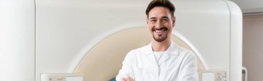 Bilgisayarlı tomografi tarayıcısı ve pankartın yanında dururken kameraya gülümseyen neşeli doktor.