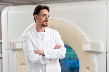 Ciddi sakallı radyoloji uzmanı klinikte tomografi cihazının yanında duruyor ve başka tarafa bakıyor.