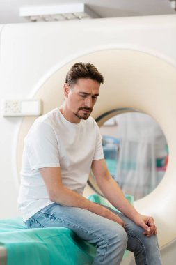 Hastanede bilgisayarlı tomografi tarayıcısının yanında oturan beyaz tişörtlü üzgün adam.