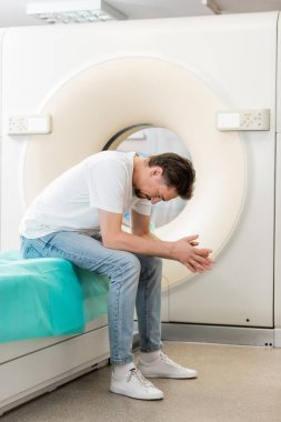 full length of depressed man sitting with bowed head near computed tomography machine clipart