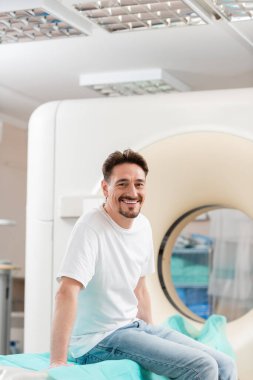 cheerful brunette man in white t-shirt sitting near ct scanner and smiling at camera clipart