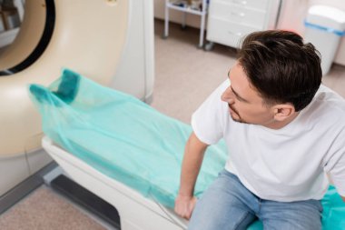 high angle view of brunette man in white t-shirt sitting near ct scanner in clinic clipart