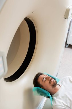 high angle view of brunette man lying during scanning procedure on computed tomography machine clipart