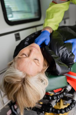 Unconscious woman lying near blurred paramedic in latex gloves in emergency vehicle  clipart