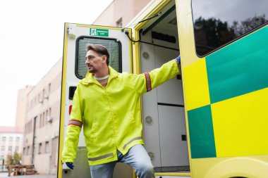 Dışarıdaki ambulans aracının kapısında duran ceketli sağlık görevlisi. 