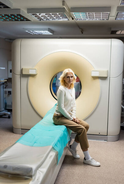 full length of blonde mature woman sitting near computed tomography machine in clinic