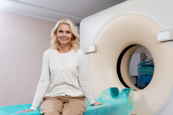 joyful blonde woman looking at camera while sitting near computed tomography scanner