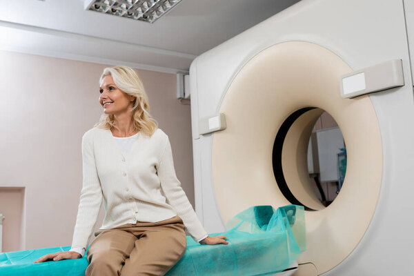 happy middle aged woman looking away while sitting near ct scanner in clinic