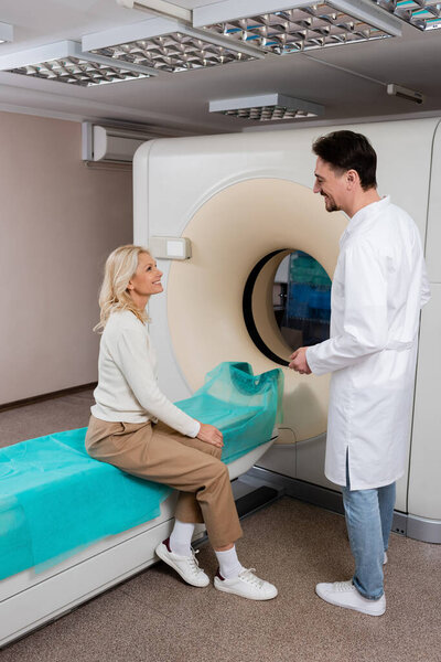 radiologist in white coat talking to smiling mature woman sitting near computed tomography machine
